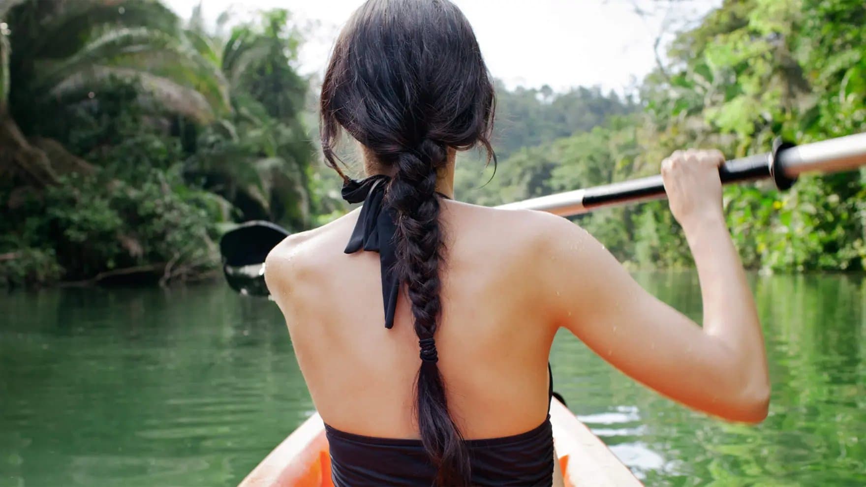 Mujer con cabello largo y negro trenzado remando en kayak por un río, de espaldas a la cámara